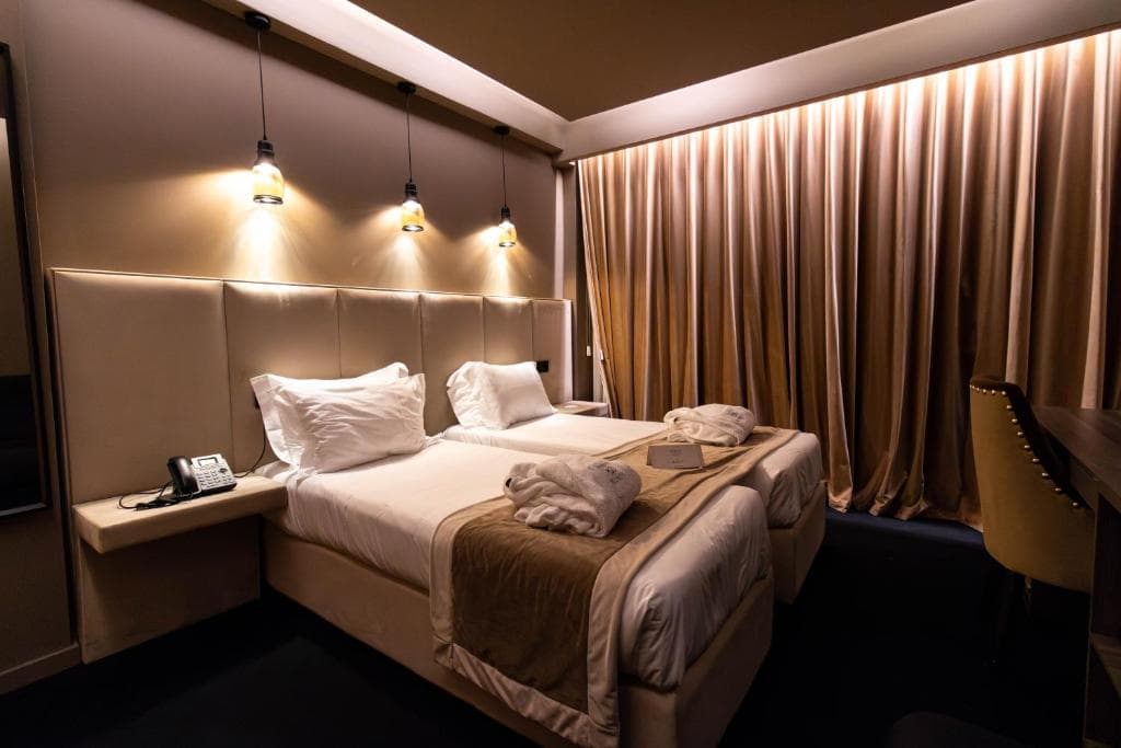 A hotel room with two neatly made single beds, white linens, beige throws, bathrobes, wall lamps above the headboard, a phone on the nightstand, and long beige curtains covering the window.