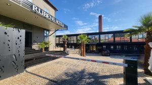 Entrance to Rubens Hotel with palm trees, a black gate arm, cobblestone pavement, and a modern black building in the background under a blue sky.