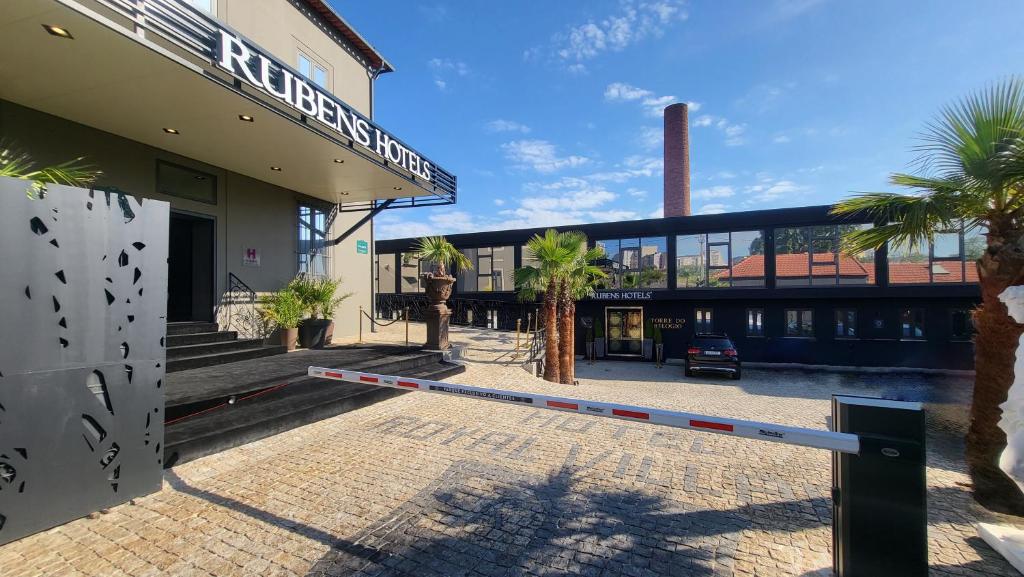 Entrance to Rubens Hotel with palm trees, a black gate arm, cobblestone pavement, and a modern black building in the background under a blue sky.