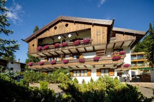 A chalet-style building with wooden balconies adorned with vibrant purple flowers, surrounded by green shrubs and trees, under a clear blue sky.