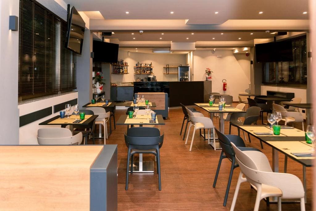 Modern restaurant interior with neatly arranged tables and chairs, place settings with glasses and green bottles, wooden floor, contemporary lighting, and a bar area with shelves of bottles in the background.