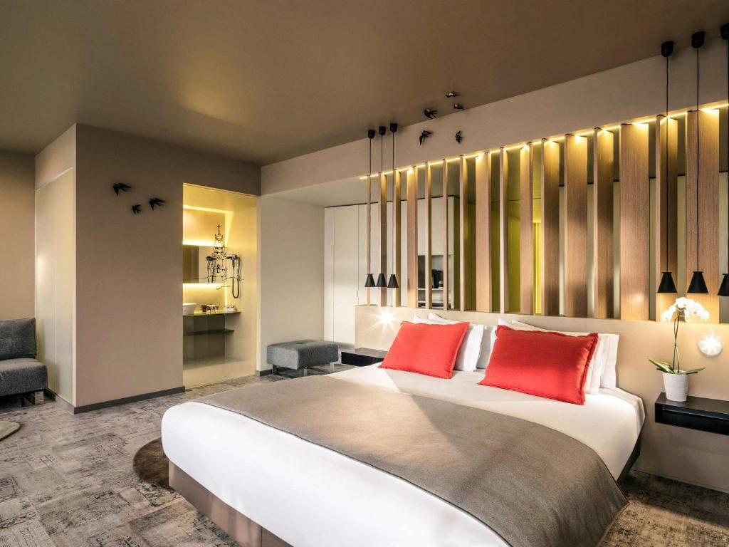 A modern hotel room with a large bed, white bedding, and two red accent pillows. Warm lighting, a wood partition, wall decor, and a bathroom area with glass shower are visible. Contemporary furnishings and neutral colors create a cozy feel.
