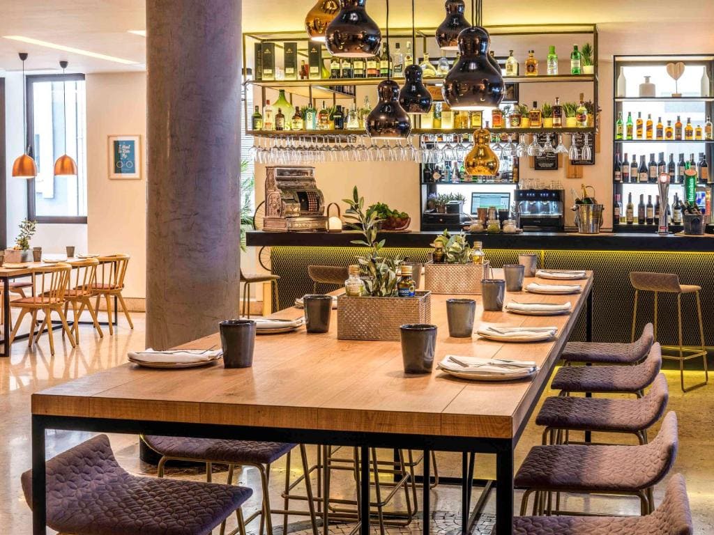 Modern restaurant interior with a large wooden dining table set with napkins and potted plants, stylish pendant lights above, bar area with bottles and glasses in the background, and additional seating by the windows.