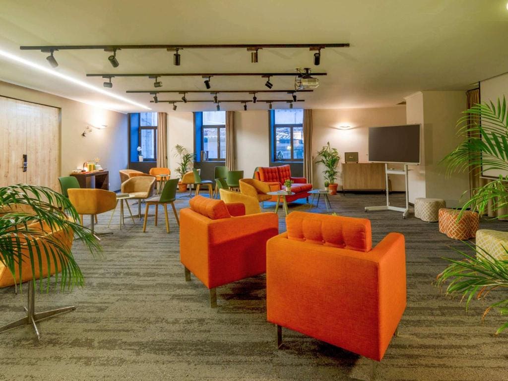 A modern lounge area with orange and yellow armchairs, small tables, green plants, and large windows. The room has warm lighting, gray carpet, and a flat-screen TV on a stand in the corner.