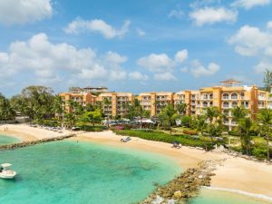 Aerial view of a tropical beachfront resort with yellow buildings, palm trees, sandy beach, turquoise water, and a small boat anchored near the shore under a blue sky with scattered clouds.