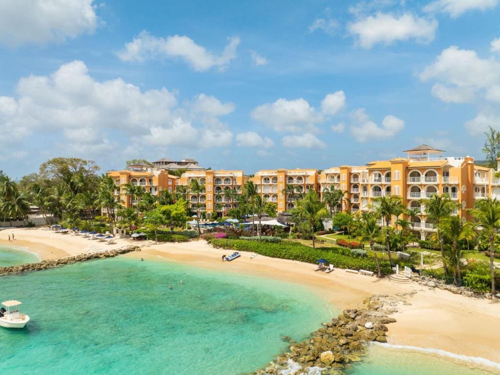 Aerial view of a tropical beachfront resort with yellow buildings, palm trees, sandy beach, turquoise water, and a small boat anchored near the shore under a blue sky with scattered clouds.