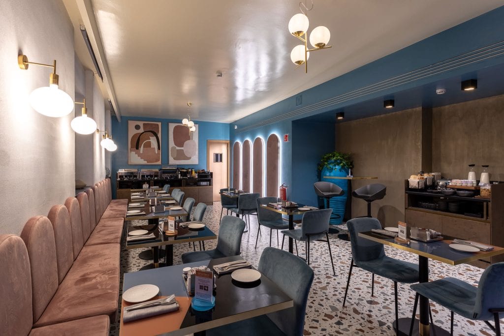 Modern restaurant interior with pink cushioned benches, blue velvet chairs, marble-topped tables, terrazzo flooring, abstract wall art, round light fixtures, and a self-serve beverage station in the corner.