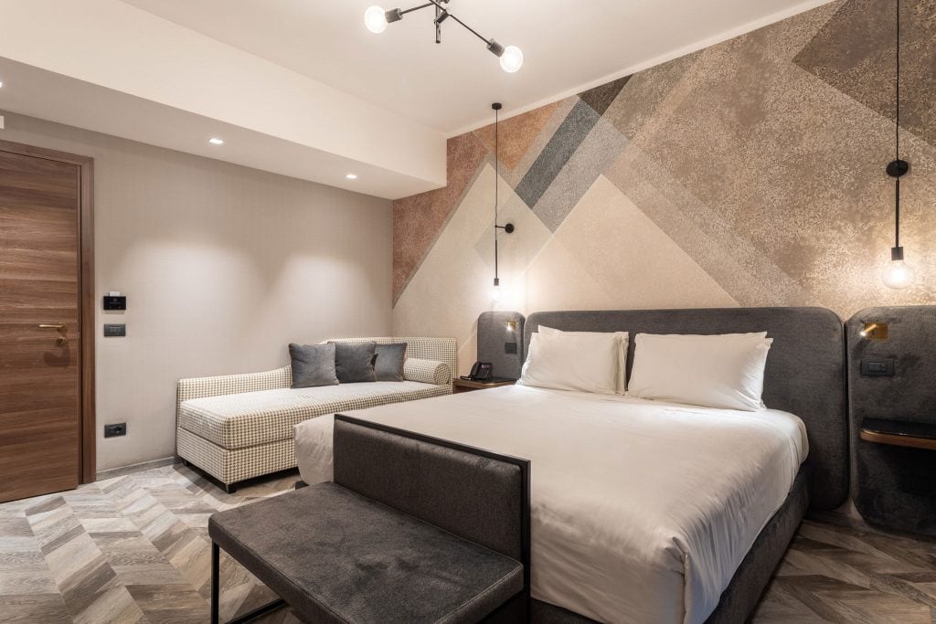 Modern hotel room with a large bed, a cushioned bench, a sofa, geometric patterned wall, pendant lights, and wood-patterned flooring. The decor is contemporary with neutral tones and minimalist furniture.