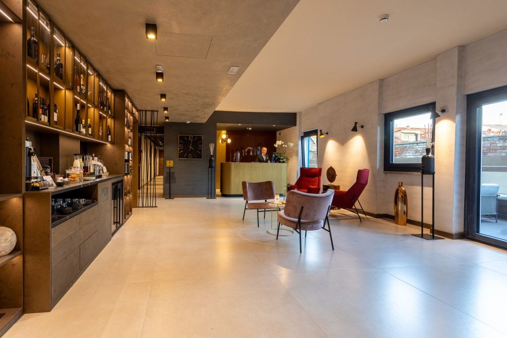 Modern lounge area with stylish armchairs, a red recliner, shelves with decor and drinks, large tile flooring, warm lighting, and windows letting in natural light. A gold reception desk is visible in the background.