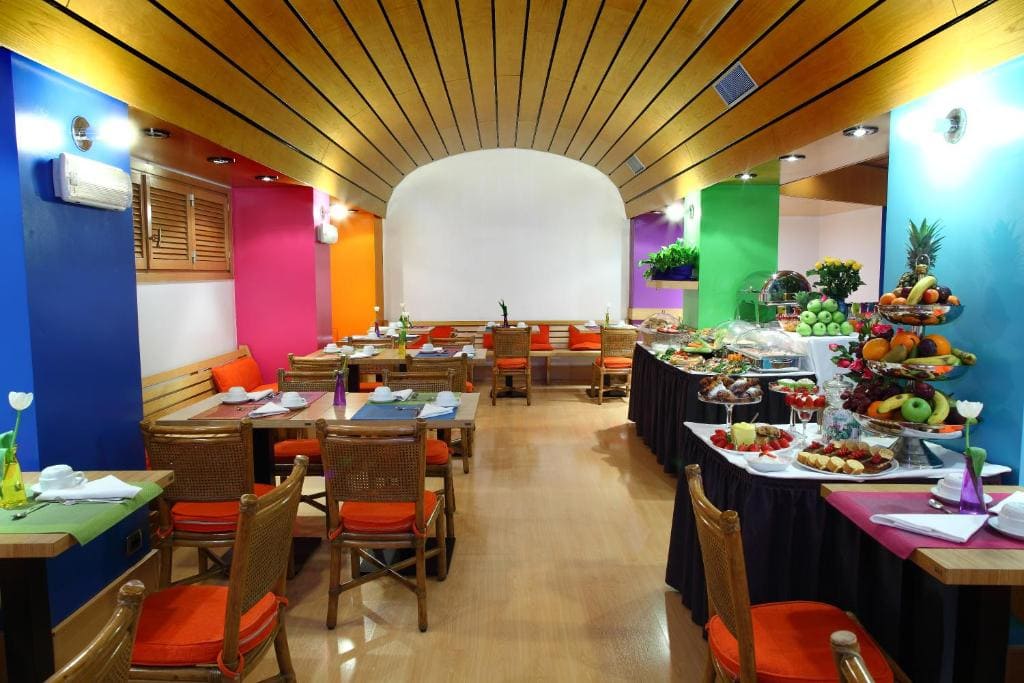 A bright, colorful restaurant with wooden chairs, orange cushions, and vibrant blue, pink, orange, and green walls. Tables are set for dining, and a buffet with various fruits and dishes is displayed on the right.