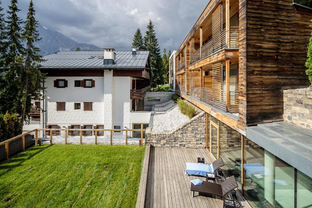 A modern wooden building with large glass windows and balconies stands beside a traditional white house with brown shutters, surrounded by green grass, a wooden deck with lounge chairs, stone walls, and tall pine trees.