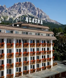 A multi-story hotel named “Alaska” with balconies decorated with red flowers, set against a backdrop of tall, rocky mountains under a clear blue sky.