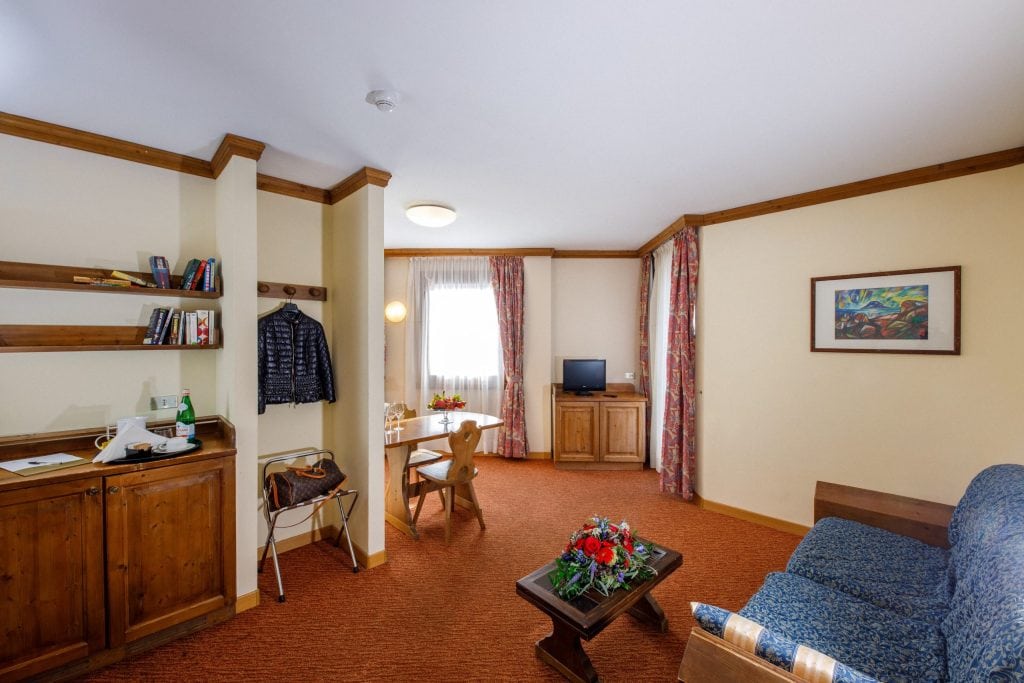 A cozy hotel room with wooden furniture, a blue patterned sofa, a desk with a chair near a window, a small TV on a cabinet, shelves with books, and a coffee table with a flower arrangement.