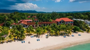Aerial view of a beachfront resort with red-roofed buildings surrounded by palm trees, blue beach chairs on the sandy shore, and clear turquoise water, with lush green hills in the background.