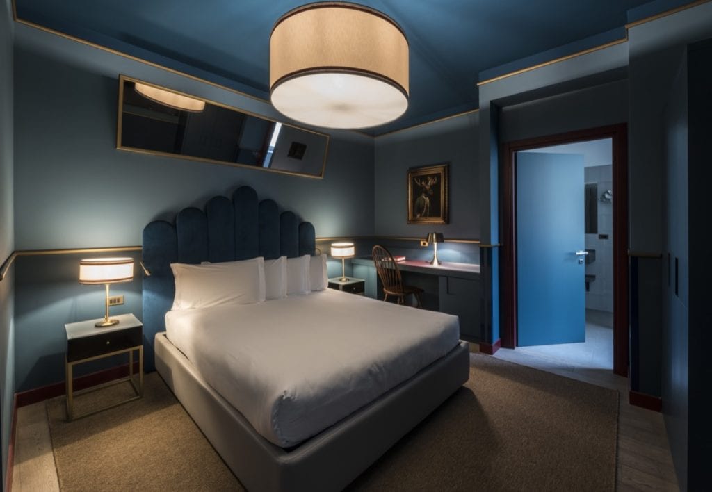 A modern hotel room with a large bed, blue upholstered headboard, two bedside tables with lamps, a desk and chair, a framed picture on the wall, and an open door leading to a bathroom.