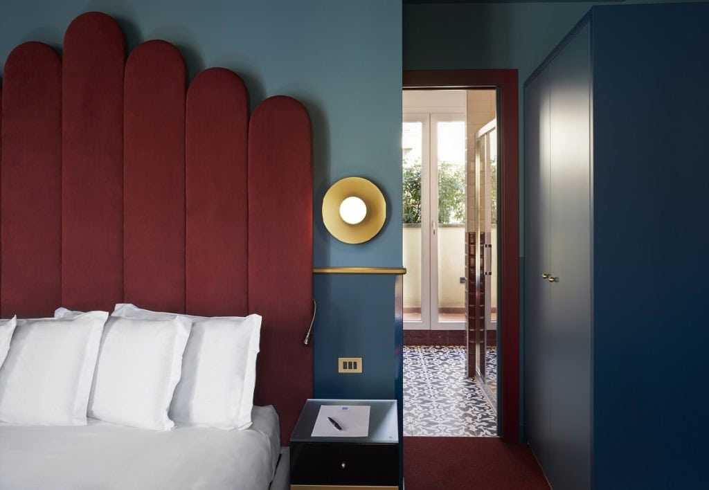 A modern bedroom with a tall, red, scalloped headboard, white pillows, a blue wall, a round gold wall light, a bedside table with a pen and notepad, and a door leading to a patterned-tile balcony.