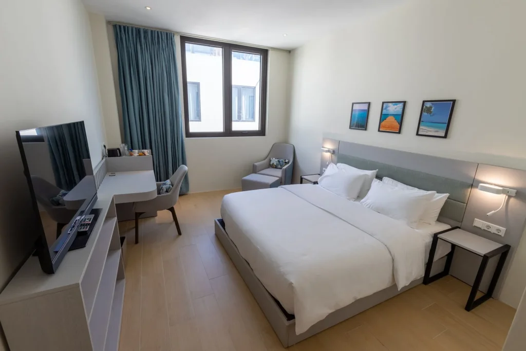 A modern hotel room with a large bed, two bedside tables, a desk with a chair, an armchair, a flat-screen TV, and three framed pictures on the wall. Light wood floors and large windows with blue curtains.
