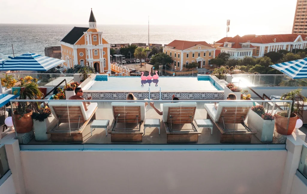 Four people relax on poolside loungers atop a rooftop, overlooking colorful buildings and the sea at sunset. Decorative flamingos are by the pool and blue striped umbrellas provide shade.