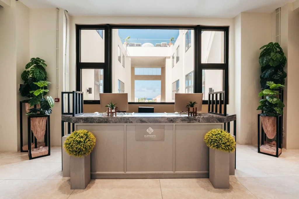 A modern reception desk with a marble top, two computers, green plants in tall pots on each side, and large windows behind letting in natural light from an inner courtyard.