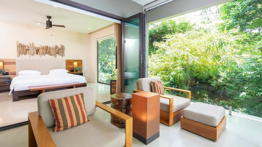 A modern hotel room with a large bed, ceiling fan, and wooden decor, connected to a bright sitting area with armchairs, ottomans, and floor-to-ceiling windows overlooking a lush green garden.