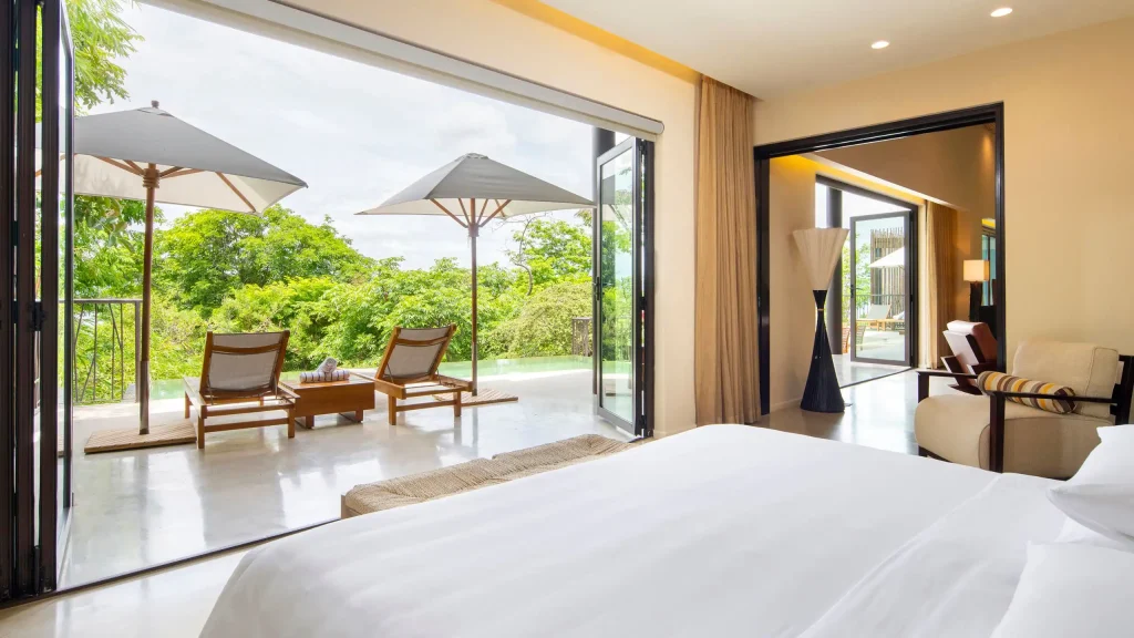 A bright, modern hotel room with a large bed, open sliding doors leading to a patio with lounge chairs, umbrellas, a small pool, and lush green trees outside under a partly cloudy sky.