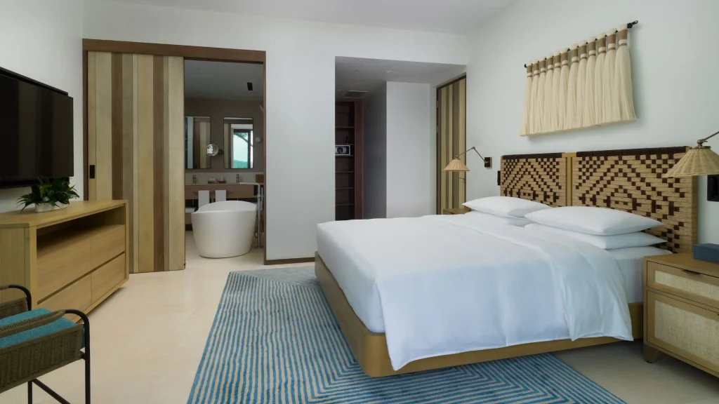A modern hotel room with a large bed, geometric headboard, blue striped rug, and light wood furniture. Sliding doors open to a bathroom with a freestanding bathtub, double sinks, and a mirror.