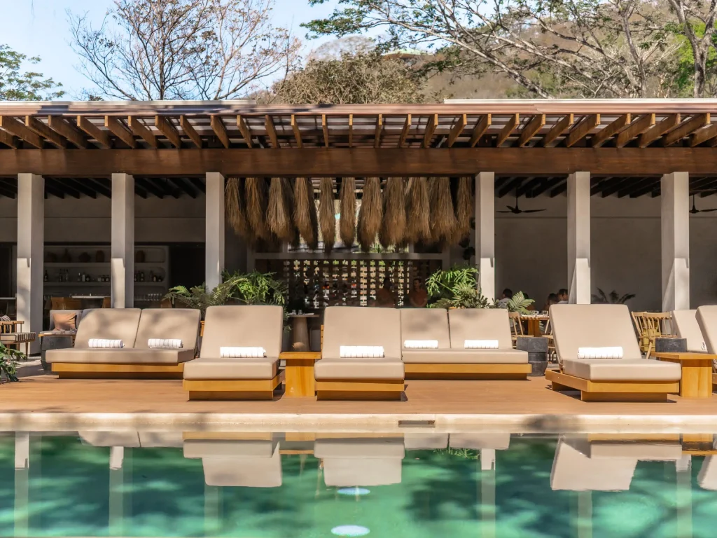 Modern outdoor lounge area with tan cushioned daybeds lined up beside a clear pool, in front of an open-air bar with hanging dried plants and surrounded by greenery and trees.