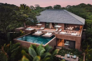 A modern luxury villa surrounded by lush greenery features a large infinity pool, sun loungers, multiple patios with seating areas, and large windows, all set in a tropical environment at sunset.