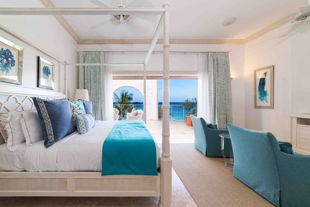 A bright, airy bedroom with a canopy bed, blue and white bedding, blue armchairs, and large glass doors opening to a balcony with ocean views and tropical plants. Coastal decor and light curtains enhance the relaxing feel.