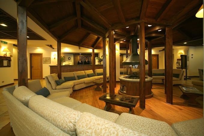 A cozy hotel lobby with wooden beams, a central fireplace, and beige sectional sofas arranged in a circle around the fireplace, creating a warm and inviting atmosphere.