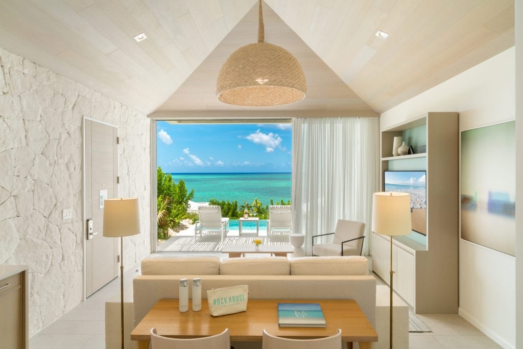 A bright, modern living room with a view of a private pool, patio, and turquoise ocean beyond. Light wood and stone decor, with large windows letting in natural sunlight and lush greenery outside.