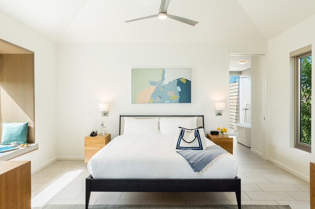 A bright, modern bedroom with a large bed, white bedding, two wooden nightstands, a ceiling fan, a window seat, abstract artwork above the bed, and an open door leading to a bathroom.