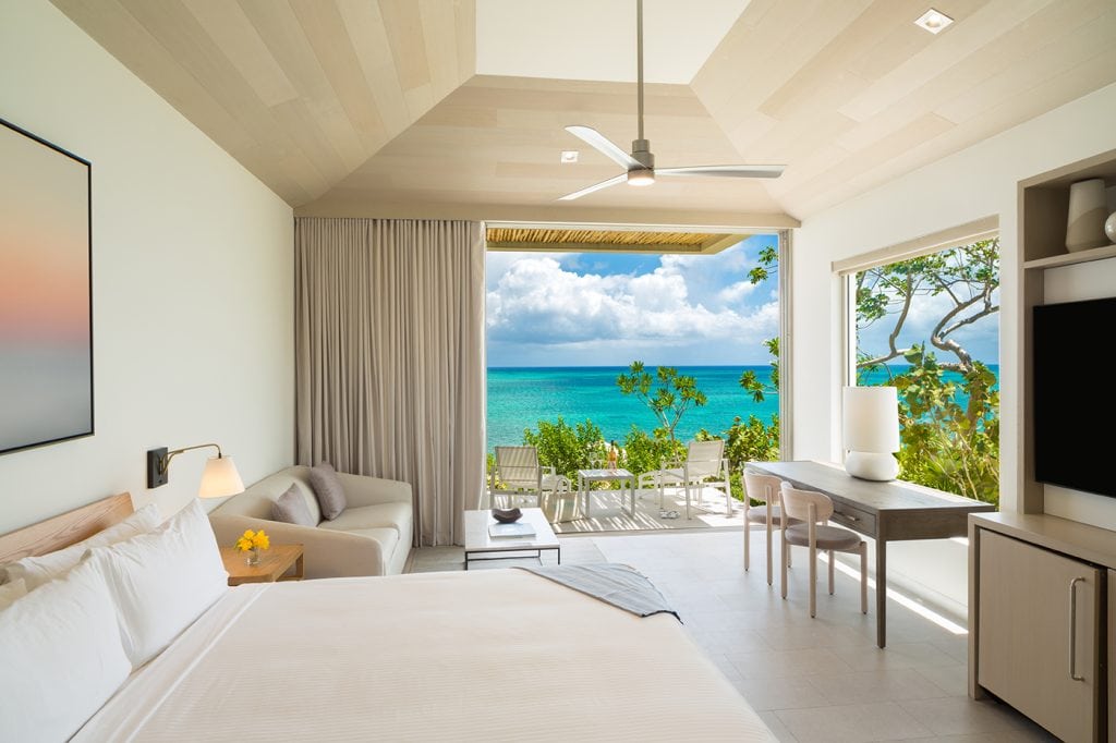 A bright, modern hotel room with a large bed, sofa, desk, and TV opens to a balcony overlooking turquoise ocean waters, blue sky, and lush greenery.