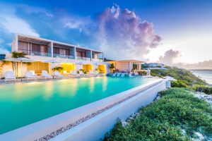 Modern luxury villa with large pool, white lounge chairs, and glowing lights at sunset. The building overlooks lush greenery and a scenic coastline with calm ocean waters under a dramatic, cloudy sky.