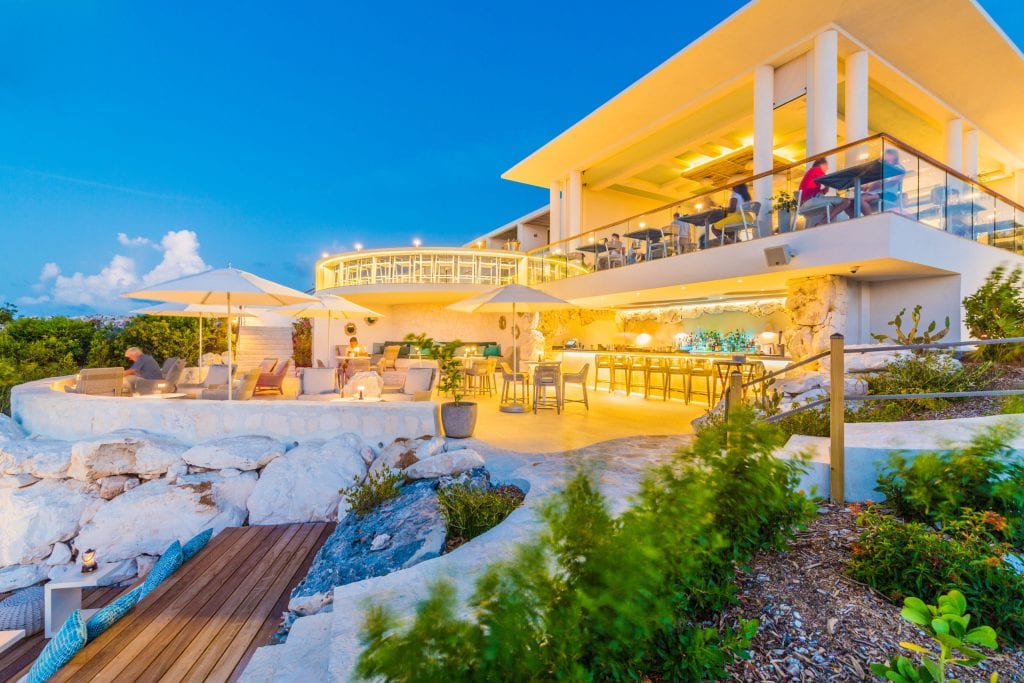 A modern, brightly-lit seaside restaurant with outdoor seating, large umbrellas, and lush greenery, overlooking the ocean at sunset. Patrons dine and relax on multiple levels of the terrace.