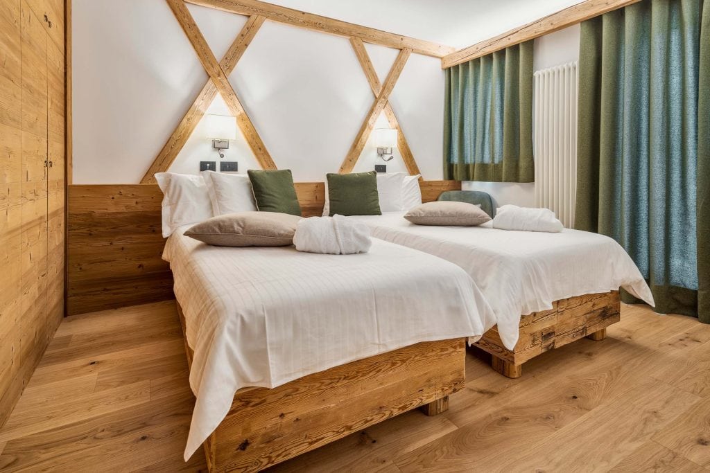 A cozy hotel room with two single beds featuring white linens, green and beige pillows, wooden headboards, and wooden floors. The room has green curtains, wall lights, and a modern, rustic design.
