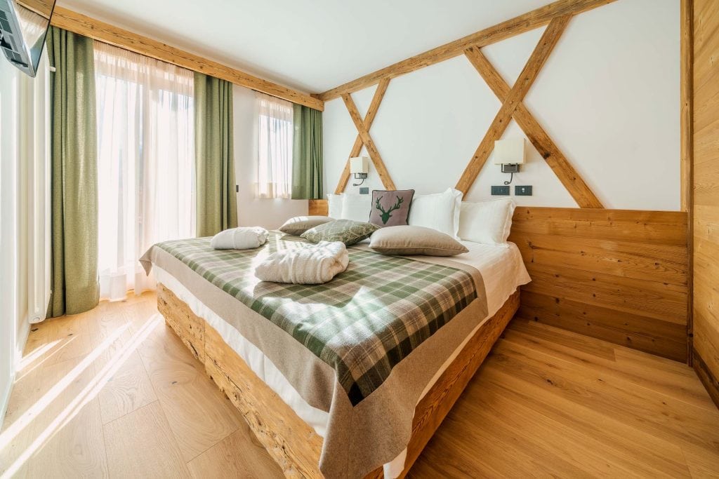 A cozy, sunlit bedroom with a large wooden bed, green plaid bedding, decorative pillows, folded robes, wood panel accents, light wood floors, and floor-to-ceiling windows with green curtains.