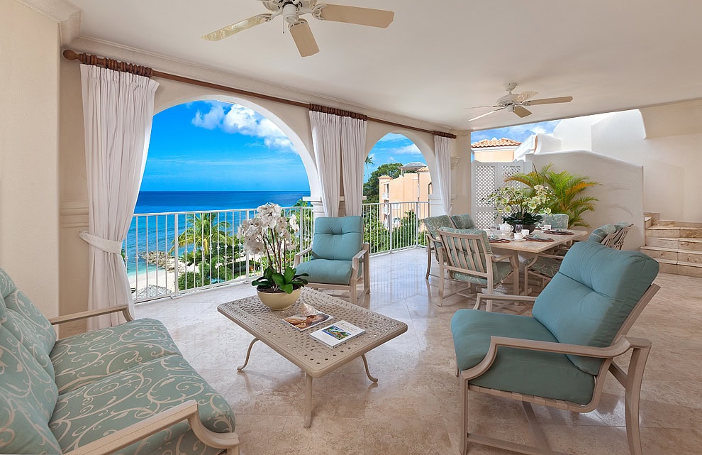 A bright, airy terrace overlooking the ocean, featuring light blue cushioned chairs, a coffee table, dining area, ceiling fans, potted plants, and large arches framing a view of palm trees and blue sky.