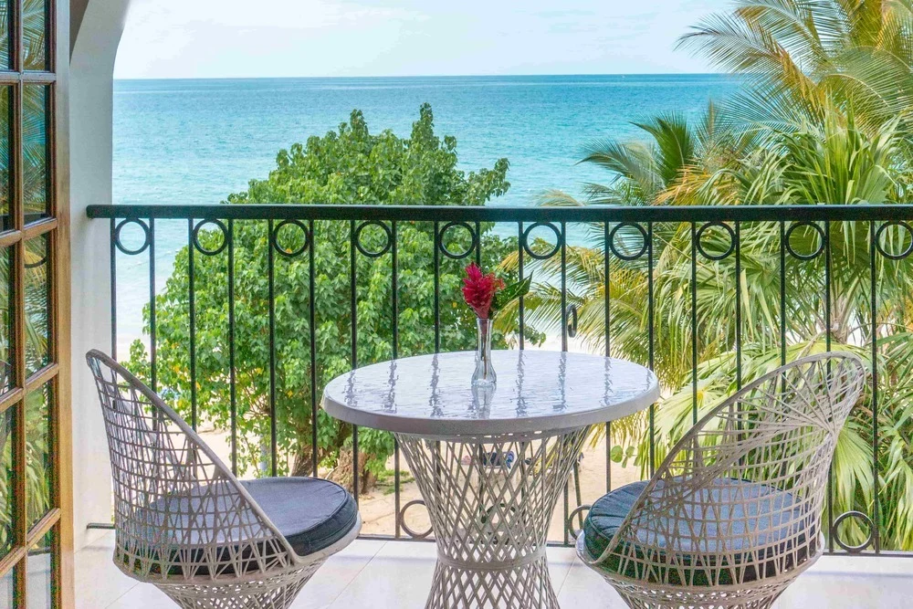 A round white table with two white chairs on a balcony overlooks tropical trees and a blue ocean under a clear sky. A small vase with a red flower sits on the table.