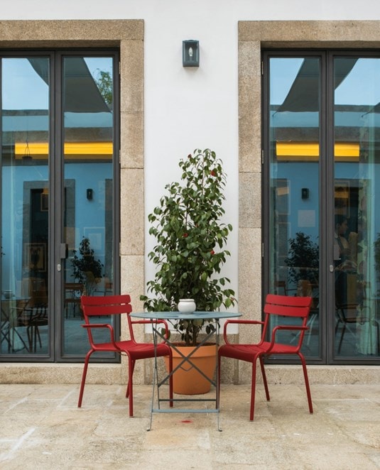 Two red chairs and a small table with a potted plant and a teapot sit on a stone patio, positioned between two tall glass doors framed by stone.