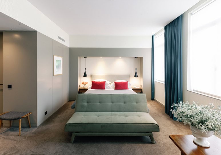 Modern hotel room with a double bed, two red pillows, a green sofa at the foot of the bed, large windows with teal curtains, a side table with white flowers, and minimalistic decor.