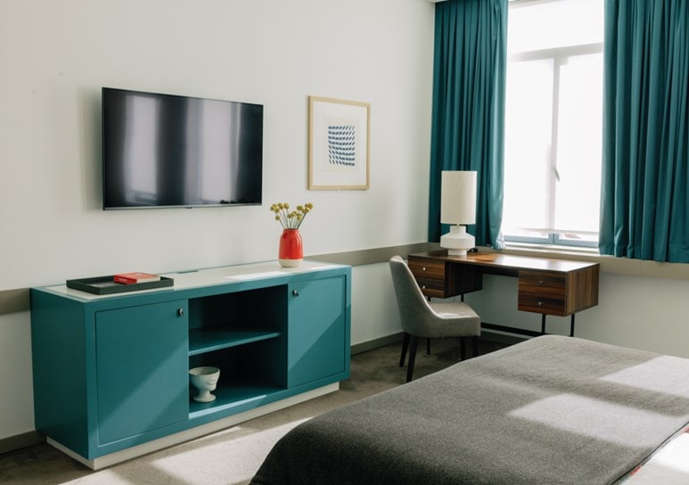 A modern, minimal bedroom with a teal cabinet, wall-mounted TV, desk with a lamp, gray chair, teal curtains, and a bed covered with a gray blanket. Light streams in from a large window.