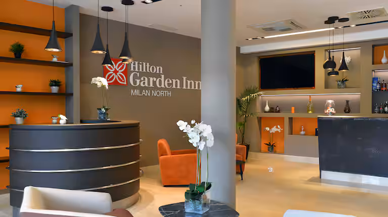 Modern hotel lobby with a round reception desk, orange chairs, potted orchids, and wall shelves. The Hilton Garden Inn Milan North logo is displayed on the wall behind the desk.