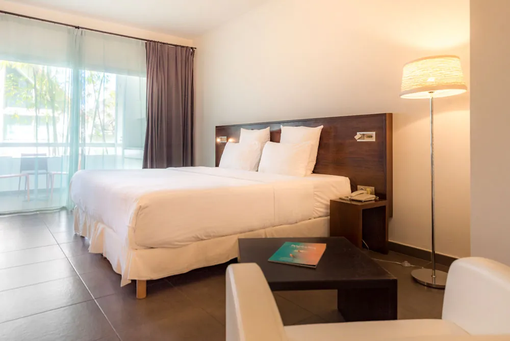 Modern hotel room with a large bed, white bedding, bedside tables, a floor lamp, a small table with a brochure, an armchair, and a window with sheer and blackout curtains letting in natural light.