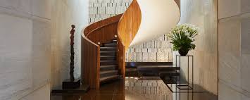 A modern interior featuring a curved wooden staircase, a tall sculpture, a large potted plant on a stand, and textured wall panels, with soft lighting creating an elegant atmosphere.