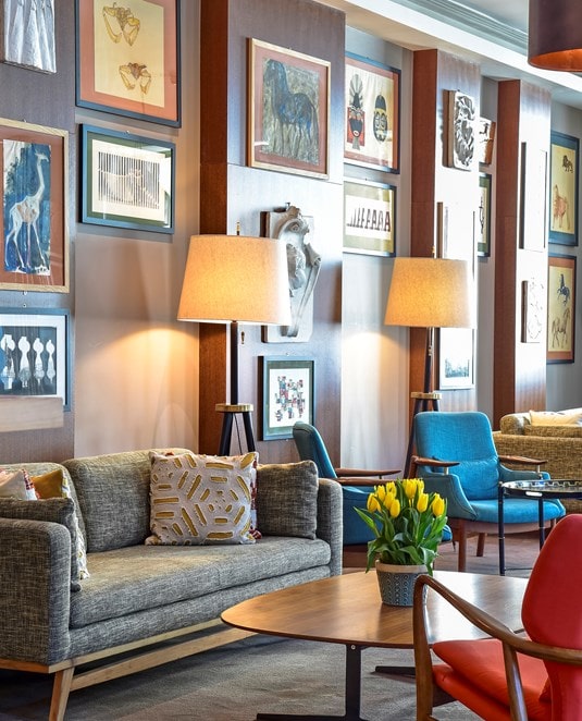 A stylish lounge with modern sofas, colorful armchairs, tall floor lamps, and a wooden coffee table with yellow tulips. The wall is decorated with an eclectic gallery of framed artwork.