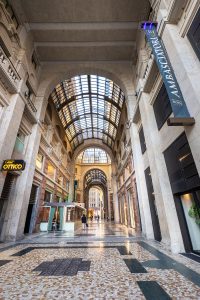 A wide, elegant shopping arcade with a high arched glass ceiling, stone columns, patterned tile floor, and storefronts along the sides. A few people walk through the bright, airy corridor.