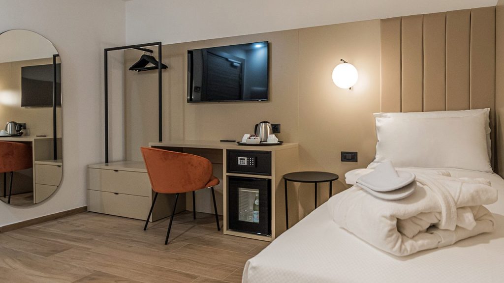 A modern hotel room with a single bed, folded towels, a desk with an orange chair, a wall-mounted TV, a mini fridge, an electric kettle, a mirror, a hanging rack, and a round wall light.