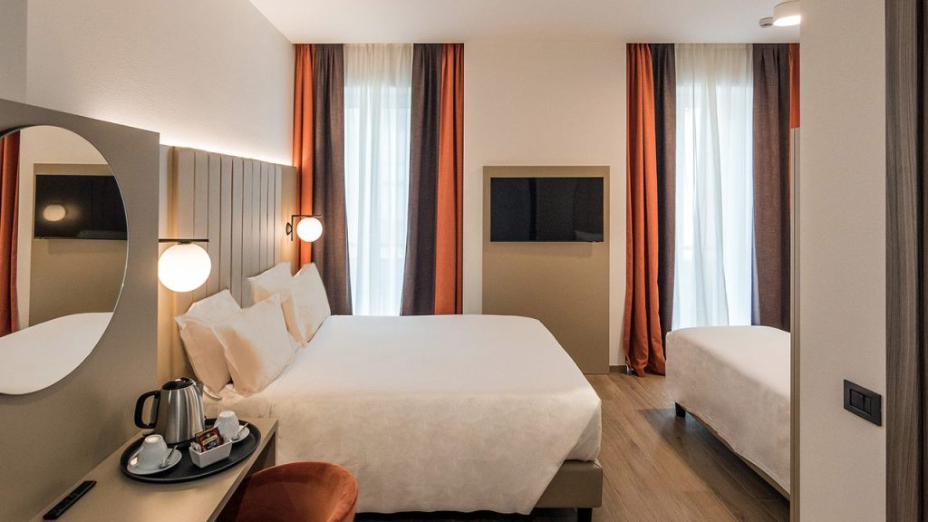Modern hotel room with a large bed, a single bed, a wall-mounted TV, tall windows with red and white curtains, and a desk with a mirror, chair, and a tray holding a kettle and cups.