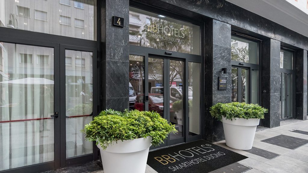 Entrance of BB Hotels Smart Hotel Duomo with glass doors, a black marble facade, large potted plants on either side of the entrance, and a logo mat on the sidewalk.